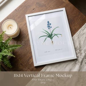 May include: A white framed botanical print featuring a blue flower illustration with green leaves. The frame is 11x14 inches. A lit candle in a glass jar and a small potted fern are on a wooden surface. The text "11x14 Vertical Frame Mockup" is at the bottom.