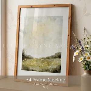 Può includere: Mockup di cornice A4 in legno chiaro con un dipinto di paesaggio. L'opera presenta un campo di fiori gialli e colline verdi sotto un cielo morbido e nuvoloso. La cornice è su una superficie di legno.