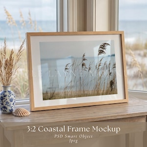 May include: A framed coastal scene mockup with a light wood frame. The image inside the frame depicts tall grasses near the ocean. A small vase with dried grasses and a seashell are on the wooden table. The text "3:2 Coastal Frame Mockup" is at the bottom.