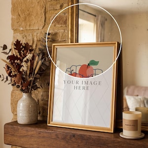 May include: A framed print with a gold border featuring a fruit illustration and the text "YOUR IMAGE HERE" sits on a wooden surface. A vase with dried flowers and a candle are also present. The scene is set against a stone wall and a mirror.