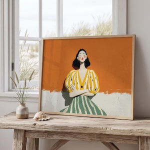 May include: A framed art print depicting a woman in a yellow and white striped top and a green and white striped skirt. The background is orange and white. The artwork is displayed on a wooden table near a window.