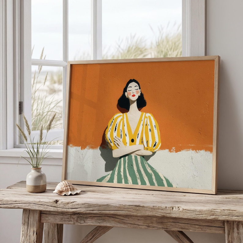 May include: A framed art print depicting a woman in a yellow and white striped top and a green and white striped skirt. The background is orange and white. The artwork is displayed on a wooden table near a window.
