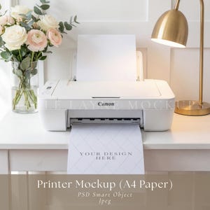 Puede incluir: Una impresora Canon blanca con una hoja de papel que dice "YOUR DESIGN HERE" está sobre una mesa blanca. Un ramo de flores en un jarrón de cristal y una lámpara de escritorio dorada también están sobre la mesa. La imagen es una maqueta de impresora.