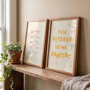 May include: Two framed art prints in wooden frames. One shows colourful fish illustrations, the other reads "THIS KITCHEN IS FOR DANCING" in yellow letters. The prints are displayed on a wooden shelf with a potted plant.