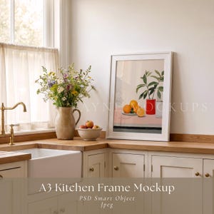 Puede incluir: Una maqueta de cocina A3 enmarcada en blanco con una pintura de naturaleza muerta de naranjas y una planta en un jarrón rojo. Un jarrón de flores y un cuenco de manzanas se encuentran sobre una encimera de madera. La cocina tiene armarios blancos y un fregadero blanco.