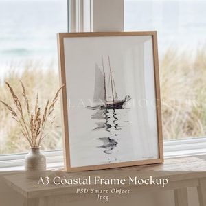 Op de afbeelding: Een ingelijste aquarelprint van een zeilboot op het water, met een licht houten frame. De print wordt weergegeven op een houten tafel bij een raam. Een vaas met gedroogde planten staat op de tafel. De tekst "A3 Coastal Frame Mockup" staat onderaan.