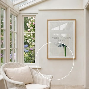 Puede incluir: Una impresi&oacute;n enmarcada con el texto "YOUR DESIGN HERE" cuelga de una pared blanca. La impresi&oacute;n tiene un marco de madera clara y presenta un dise&ntilde;o floral. Una silla de mimbre blanca con un coj&iacute;n color crema est&aacute; frente a una ventana.