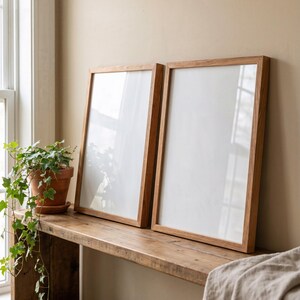 May include: Two rectangular wooden picture frames with white interiors. The frames are made of light brown wood and lean against a beige wall. A potted plant sits on a wooden shelf to the left.