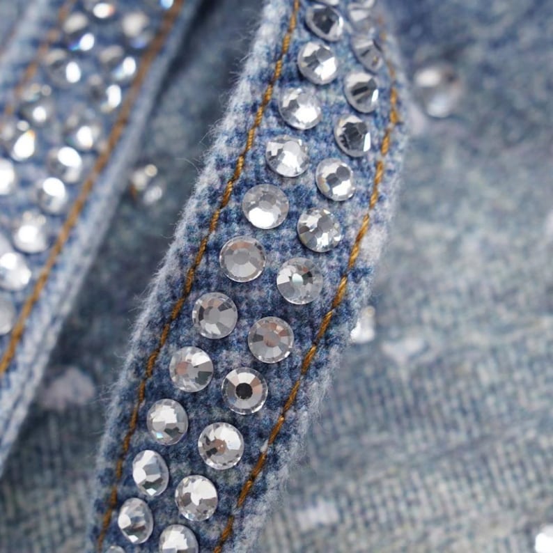 May include: Close-up of a denim fabric with sparkling, clear rhinestones. The blue fabric is accented with gold stitching. The rhinestones are evenly spaced and add a touch of glamour to the denim.