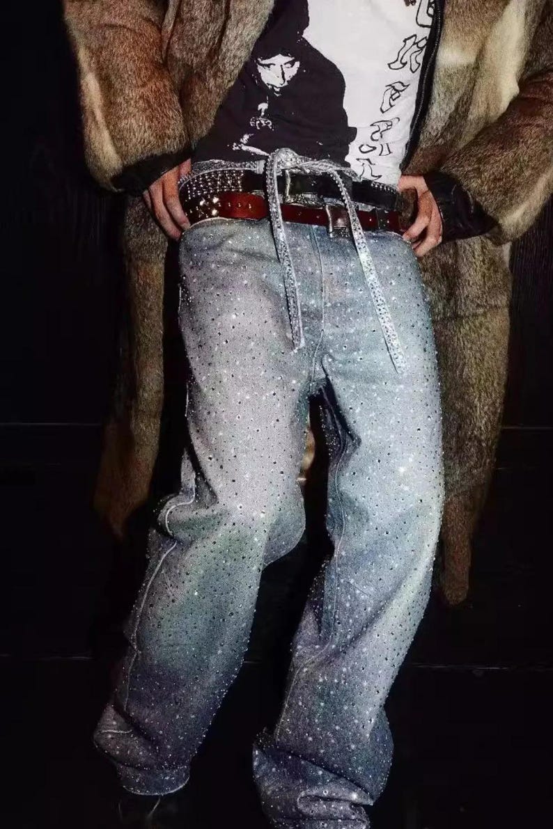 May include: Light blue denim jeans heavily embellished with sparkling rhinestones. The jeans are paired with a black and white graphic tee, a studded belt, and a brown leather belt. A brown fur coat is worn over the outfit.