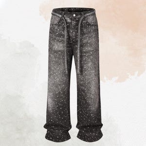 May include: Black denim jeans with a white speckled pattern. The jeans have a drawstring waist and a button closure. The pockets and waistline are embellished with a silver-colored trim. The jeans have a wide-leg fit.