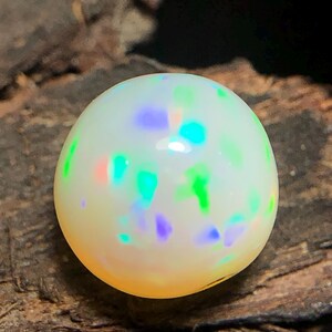 Könnte beinhalten: Ein runder, polierter Opal-Edelstein mit cremeweißer Grundfarbe. Der Opal weist eine Streuung von leuchtenden, irisierenden Farben auf, darunter Grün, Blau und Lila. Der Edelstein liegt auf einer dunklen, strukturierten Holzoberfläche.