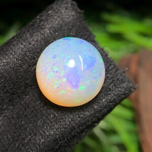 Könnte beinhalten: Ein runder, polierter Opal-Edelstein mit milchig-weißer Grundfarbe. Der Opal zeigt irisierende Blitze in Blau, Grün und Lila auf seiner Oberfläche. Der Edelstein liegt auf einem dunklen, strukturierten Hintergrund. Durchmesser: ca. 1 cm.