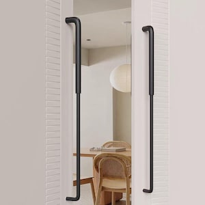 Può includere: Maniglie per porte in metallo nero con parte superiore e inferiore curve, installate su un telaio di porta bianco. Le maniglie sono lunghe e verticali, con un design minimalista. Lo sfondo mostra una zona pranzo con tavolo e sedie in legno.