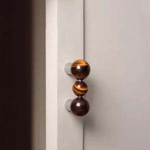 May include: A set of three decorative cabinet knobs. Each knob is a polished sphere of tiger's eye gemstone, displaying bands of brown and gold. The knobs are mounted on small, round, silver-colored bases, attached to a light gray cabinet door.