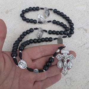 Puede incluir: Rosario de cuentas negras con crucifijo y medallones religiosos plateados. El crucifijo presenta una representación de Jesús y la inscripción "JESUS NAZARENUS REX JUDAEORUM". El rosario se sostiene en una mano.