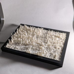 Op de afbeelding: Een 3D-geprint model van een stad, met talrijke witte gebouwen van verschillende hoogtes, geplaatst in een zwart rechthoekig frame. Het model toont een gedetailleerd stadslandschap, met straten en structuren.