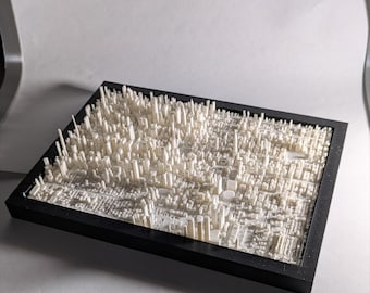 May include: A 3D-printed model of a city, featuring numerous white buildings of varying heights, set within a black rectangular frame. The model showcases a detailed urban landscape, with streets and structures.