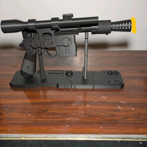 May include: A matte black prop blaster model featuring a long top-mounted scope, a textured grip, and a perforated barrel tip with a yellow accent. The blaster is mounted on a rectangular black display stand with geometric detailing and support struts.