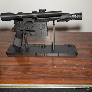 May include: A black replica blaster pistol, a prop from a science fiction film, is displayed on a black stand. The pistol has a scope, a barrel with holes, and intricate details. The stand has support rods and a rectangular base.