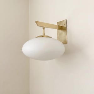 Puede incluir: Aplique de pared moderno con acabado en latón. Cuenta con una placa de pared rectangular, un brazo horizontal y una pantalla de vidrio blanco de forma ovalada que apunta hacia abajo. Montado en una pared de color neutro.