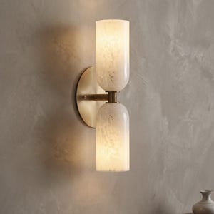 Può includere: Applique a doppia luce con due paralumi cilindrici in vetro testurizzato bianco sporco. L'applique ha una base in metallo color ottone e una piastra rotonda. Il design è montato su una parete grigia. L'applique è etichettata "THE AURORA DOUBLE SCONCE DESIGN: L. HOFFMAN (2024)".