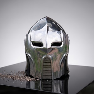 May include: A polished silver-coloured metal helmet with a pointed crown, two horizontal eye openings, and a central vertical grill section covering the mouth area. The helmet features a smooth, reflective surface with curved, moulded contours.