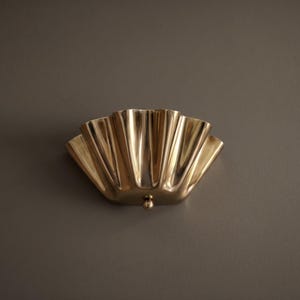 May include: A brass-colored wall sconce with a fan-shaped design. The sconce has a series of vertical ridges and a small, round detail at the bottom. The fixture is mounted on a neutral-colored wall.