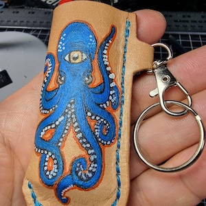 Puede incluir: Una funda de cuero color canela para encendedor con un diseño de pulpo azul y detalles en naranja. El pulpo tiene un solo ojo y puntos blancos. La funda tiene un borde cosido azul y un llavero plateado.