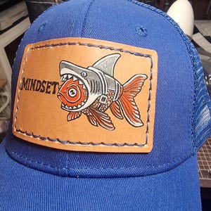 May include: A royal blue trucker hat featuring a rectangular tan leather patch stitched onto the front. The patch displays a graphic of a shark wearing a harness over a goldfish, with the word MINDSET printed in black lettering to the left.