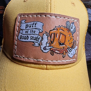 May include: A bright yellow baseball cap featuring a rectangular tan leather patch stitched to the front. The patch displays a hand-painted orange pufferfish smoking, with a banner containing the text "puff of the good stuff" in black ink.