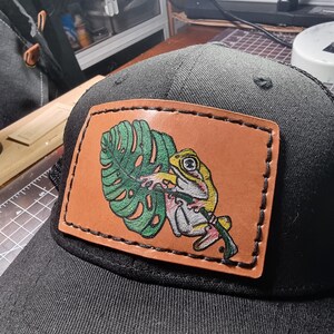 May include: A black baseball cap featuring a rectangular tan leather patch stitched onto the front. The patch displays a detailed illustration of a yellow and white frog clinging to a large, textured green monstera leaf with black outlines.