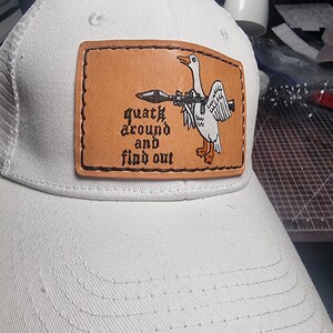May include: A white baseball cap featuring a rectangular tan leather patch stitched to the front. The patch displays a graphic of a white goose holding a rocket launcher with the black text "quack around and find out" printed to the left.