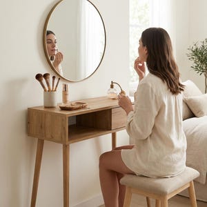 Op de afbeelding: Een vrouw brengt make-up aan bij een houten kaptafel met een ronde, goudomrande spiegel. De kaptafel heeft een lade en een open plank, met make-up borstels, een foundationfles en een klein schaaltje. Er staat een krukje voor.