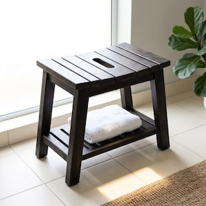 May include: A dark brown wooden stool with a slatted top featuring a central oval cutout handle. The stool has four sturdy legs and a lower slatted shelf that holds a folded white towel. The design is rectangular with clean, straight lines.