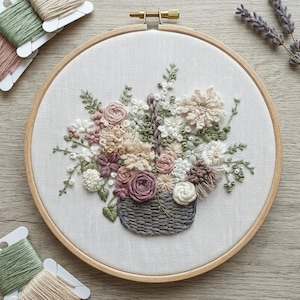 以下が含まれることがあります： 白い布に手刺繍された花のバスケットのデザイン。木製の刺繍枠に収められています。バスケットはピンク、クリーム、緑の色合いの花でいっぱいです。お揃いの糸巻きとラベンダーの小枝が枠の周りに配置されています。