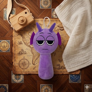 Puede incluir: Un peluche de alienígena morado con ojos grandes y somnolientos y cuernos pequeños. El juguete tiene una expresión aburrida y un lazo blanco para colgar. El fondo incluye una cámara de juguete de madera, un mapa y una tela blanca.