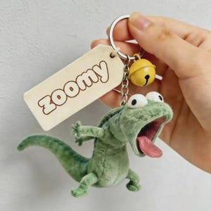 Pode incluir: Um chaveiro de lagarto verde de pel&uacute;cia com olhos grandes e boca aberta, revelando uma l&iacute;ngua rosa. O chaveiro inclui um pequeno sino amarelo e uma etiqueta que diz "zoomy".