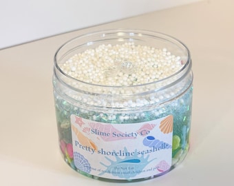 May include: A clear plastic jar filled with white foam balls, blue and green glitter, and colorful small stones. The jar has a label that reads "Slime Society Co Pretty shoreline seashell".