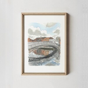 May include: Watercolor painting of a bridge over a river, framed in a gold-colored frame. The bridge is white with a curved design, and the buildings on the riverbank are a mix of orange and white. The sky is blue with fluffy clouds.