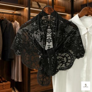 Puede incluir: Un chal de encaje negro con un estampado floral se exhibe en una percha de madera. La delicada tela está drapeada sobre una camisa blanca, mostrando el intrincado diseño y la textura. Los bordes del chal son festoneados, añadiendo un toque de elegancia.