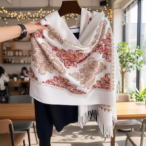 May include: A cream-coloured scarf featuring a paisley design in brown, red, and yellow. The scarf has a fringed edge and is draped over a black garment. The scarf is being held up by a person.