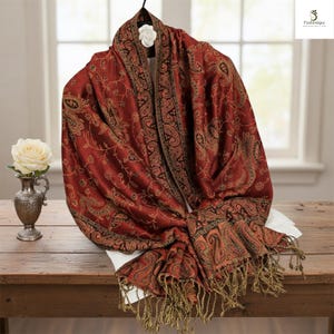 May include: A red paisley shawl with a brown border and fringe. The shawl is draped over a hanger and displayed on a wooden table. A white rose in a silver vase is on the left.