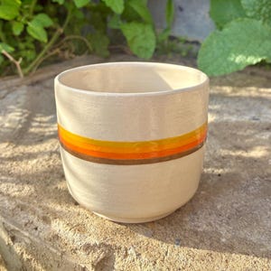 May include: A cream-coloured ceramic cup featuring a horizontal band of three distinct stripes in yellow, orange, and brown. The cup has a smooth, cylindrical shape with a slightly tapered base and a wide, open top, showcasing a clean finish.