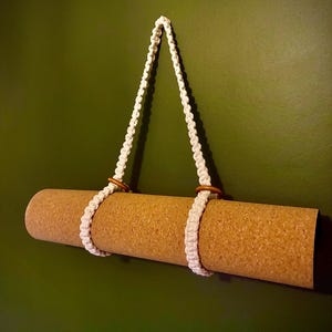 May include: A rolled cork yoga mat suspended by a white macrame holder with wooden rings. The holder is attached to a wall. The mat is a light brown color, and the wall is a dark green.
