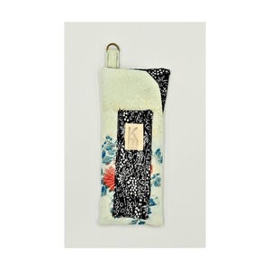 May include: A rectangular fabric case in pale green featuring a floral pattern with red and blue accents. A vertical black strip with a white botanical print runs down the centre, topped with a small rectangular tag displaying the letters KM.