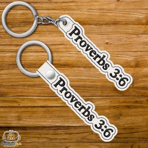 Può includere: Due portachiavi con anelli argentati ed etichette bianche. Le etichette riportano la scritta "Proverbi 3:6" in nero. I portachiavi sono su una superficie di legno.