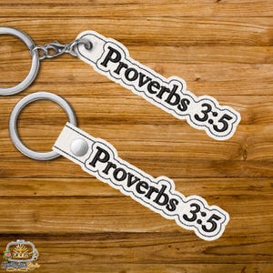 Può includere: Due portachiavi bianchi con la scritta "Proverbi 3:5" in nero. I portachiavi hanno un portachiavi argentato e una chiusura a scatto. Lo sfondo è una superficie di legno.