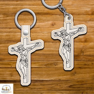 May include: Two keychains featuring a cross with a depiction of Jesus Christ. The cross and figure are outlined in black stitching on a white background, with a gold-colored border. Each keychain has a silver-colored ring.