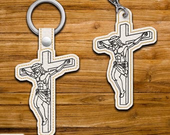 Puede incluir: Dos llaveros con una cruz con una representación de Jesucristo. La cruz y la figura están delineadas con costuras negras sobre un fondo blanco, con un borde dorado. Cada llavero tiene un anillo plateado.